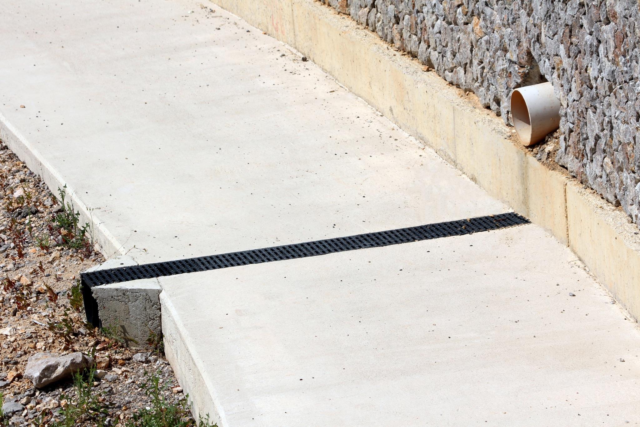 Drainage outlet pipe exiting the base of a stone retaining wall beside a concrete walkway with a channel drain