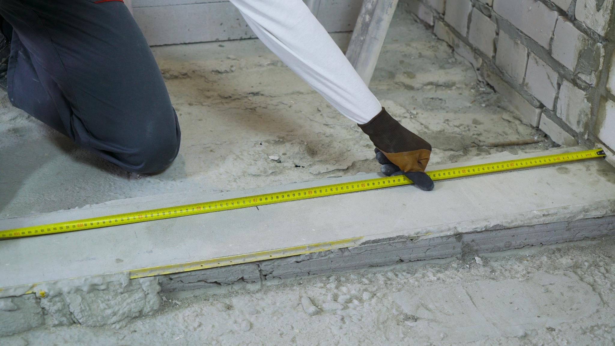 Mason kneeling to measure a concrete wall footing with a tape measure during base preparation